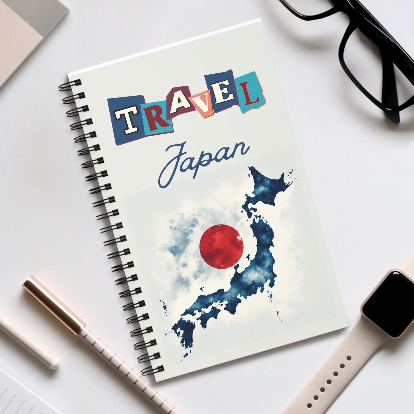 A Travel Japan Notebook by Prodigi, with a spiral binding and a Travel Japan cover featuring a map of Japan overlaid with a red circle, rests on white. Nearby are black glasses, smartwatch, pen, and closed laptop—ideal for travel journaling adventures.