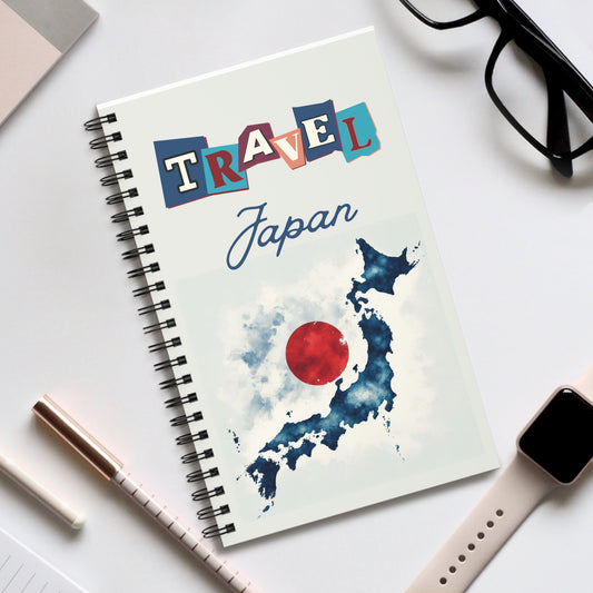 A Travel Japan Notebook by Prodigi, with a spiral binding and a Travel Japan cover featuring a map of Japan overlaid with a red circle, rests on white. Nearby are black glasses, smartwatch, pen, and closed laptop—ideal for travel journaling adventures.