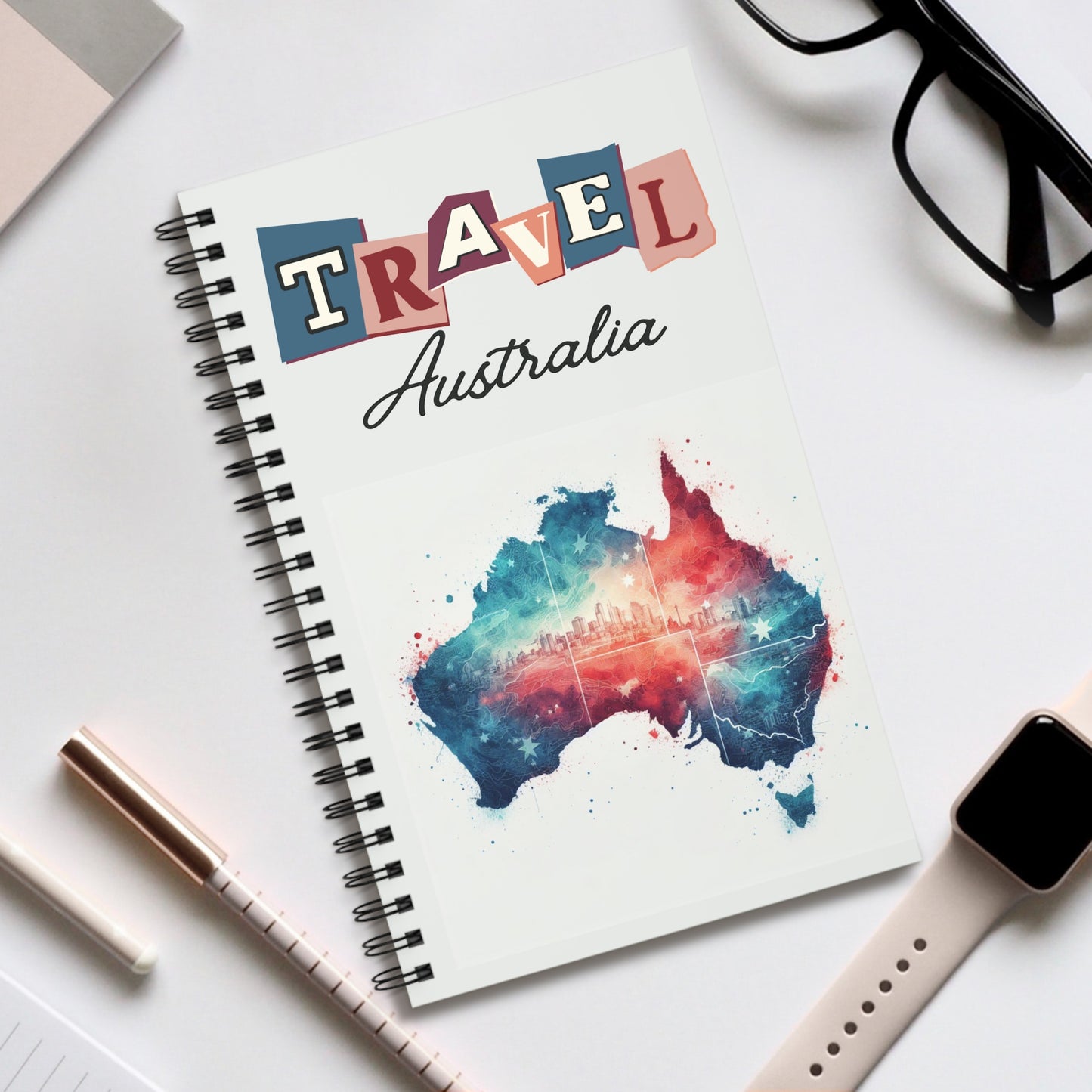 The Travel Australia spiral-bound notebook by Prodigi, featuring a colorful map and cityscape, rests on the desk. Nearby are glasses, a pen, and a smartwatch with a beige strap—ideal for jotting down adventures in this Australian travel journal with your choice of dot grid, lined or blank pages.