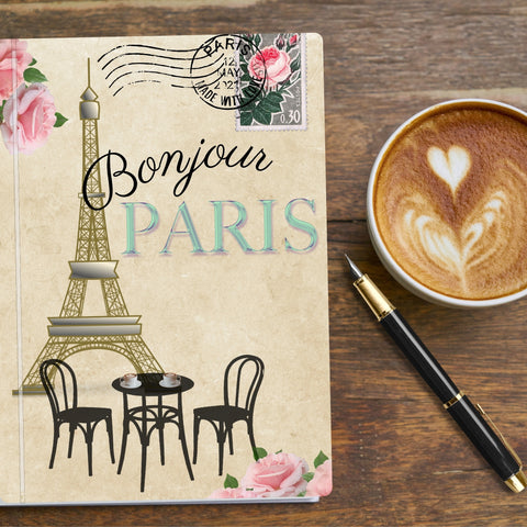 Bonjour Paris travel journal and planner  on table with a cup of coffee and pen.  Cover features eiffel tower, cafe table and chairs, french postage stamp and gloral decoration