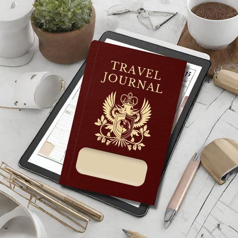 travel journal looking like a burgundy red passport with blank window for personalisation.  Shown on a table with various pens, glasses and plants