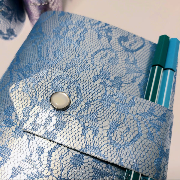 close up of the blue lace design  leather with strap fastening with a pearly snap fastener.  2 pens are tucked into the strap which is acting as a pen holderein