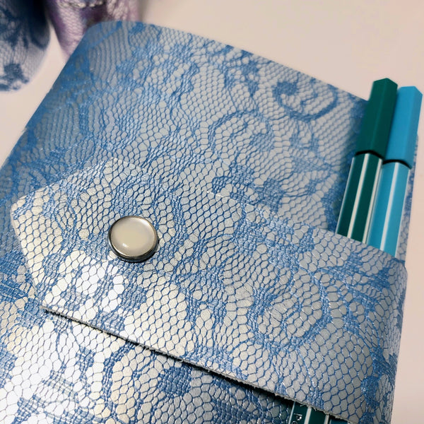 close up of the lacey design on the leather cover  of a mixed paper journal.  A green and blue pen are tucked behind the strap, acting as a pen loop