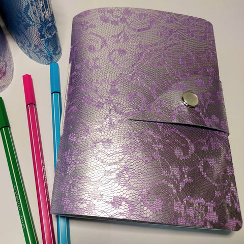 purple leather mixed paper journal closed with strap and pearly snap fastener.  3 pens and 2 further journals can be seen