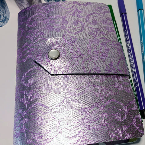 lilac/purple A6 journal in embossed leather.  A lace design with a strap and pearly snap fastener.  3 pens alongside