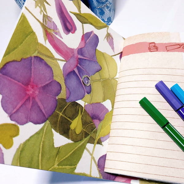 inside cover and first page of a leather mixed paper journal.  Exotic flowers decorate the cover with a lined page facing.  colourful pens lie on the page