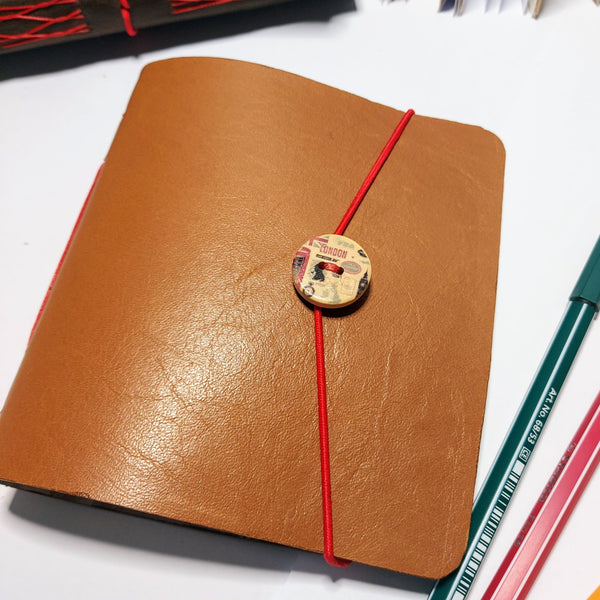 Goden brown leather cover on assorted paper journal with red elastic fastening around a large london button