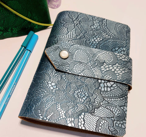 petrol blue lace design journal with pearly stud strap fastening.   tow pens alongside