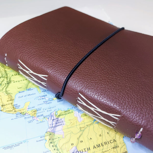 Textured brown leather travel journal with elastic loop closure showing spine with french link stitch binding and 2 small glass beads