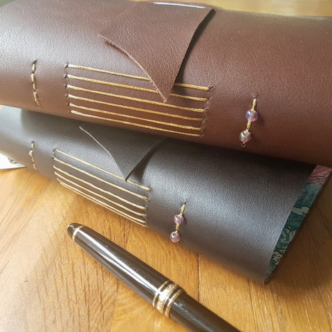 2 leather travel journals in chocolate and dark brown showing hand stitching and glass beads on spine
